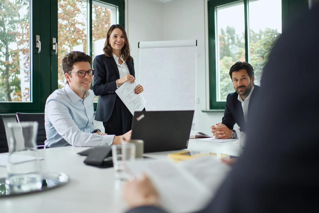 Three business people in a modern office: a woman with documents at a flip chart, two men at a table with a laptop.