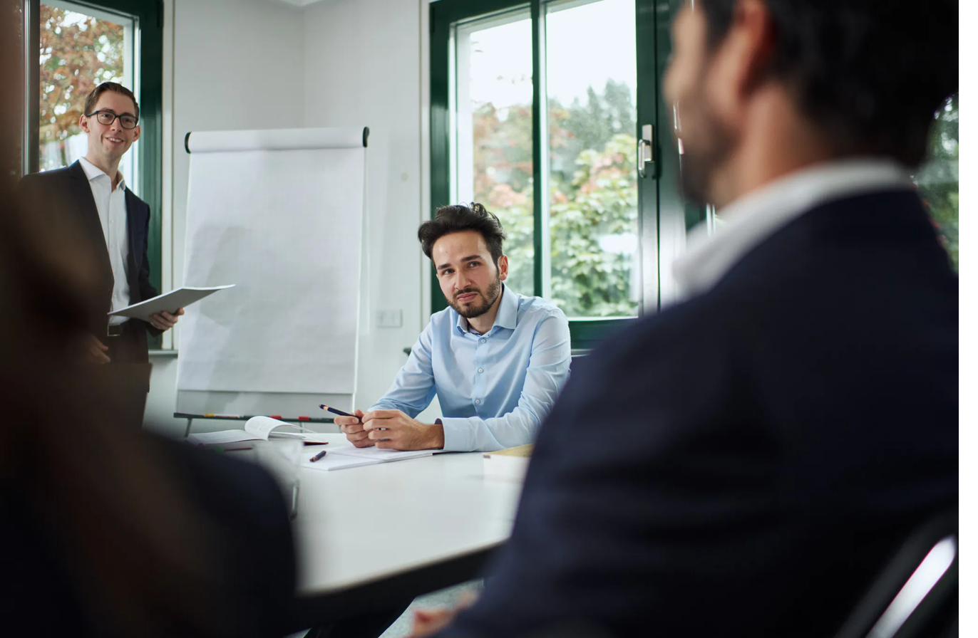 A man in a blue shirt listening attentively in a meeting with a colleague standing at a flip chart.